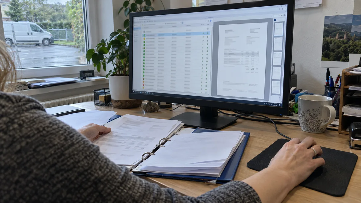 Účtovníčka v kancelárii kontroluje na monitore zoznam faktúr a náhľad dokladu, vedľa leží šanón s papierovými faktúrami.