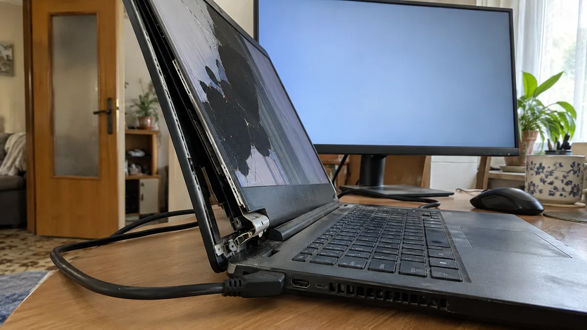 Notebook s prasknutým displejom a uvoľneným rámom pri kĺbe, pripojený káblom HDMI k externému monitoru na stole.