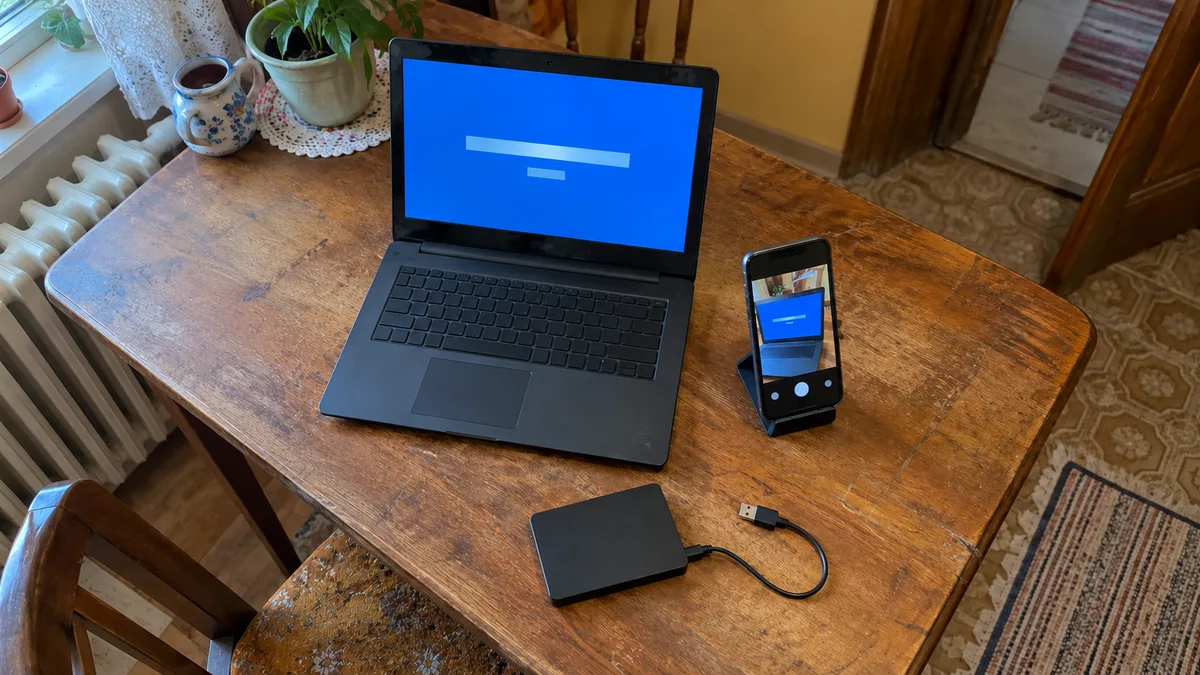 Notebook s modrou prihlasovacou obrazovkou na drevenom stole, mobil v stojane, ktorý obrazovku fotí, a externý disk s USB káblom.